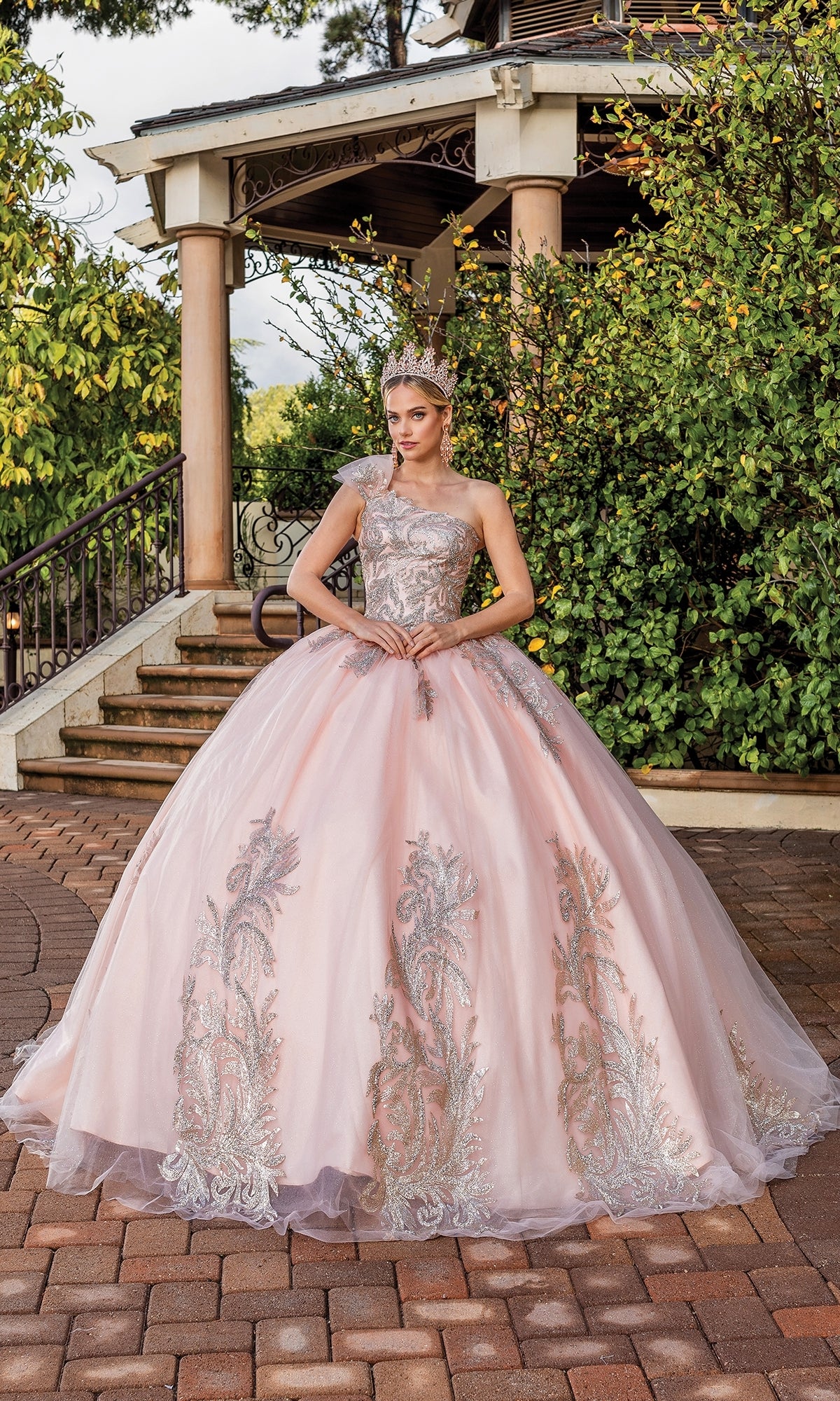 A woman in the Dancing Queen 1733 one-shoulder long quince dress stands on a brick path before a garden gazebo.